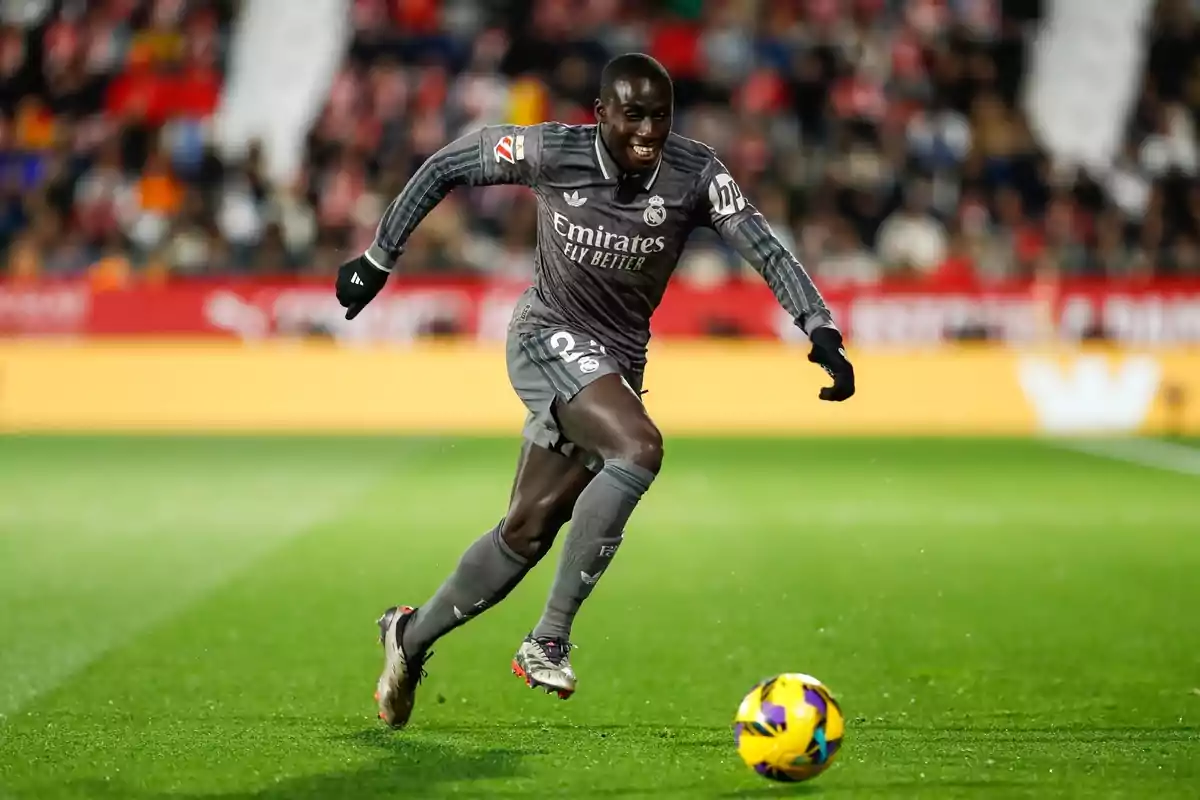 Un jugador de fútbol con uniforme gris corre hacia un balón en el campo durante un partido. Un jugador de fútbol con uniforme gris corre hacia un balón en el campo durante un partido.