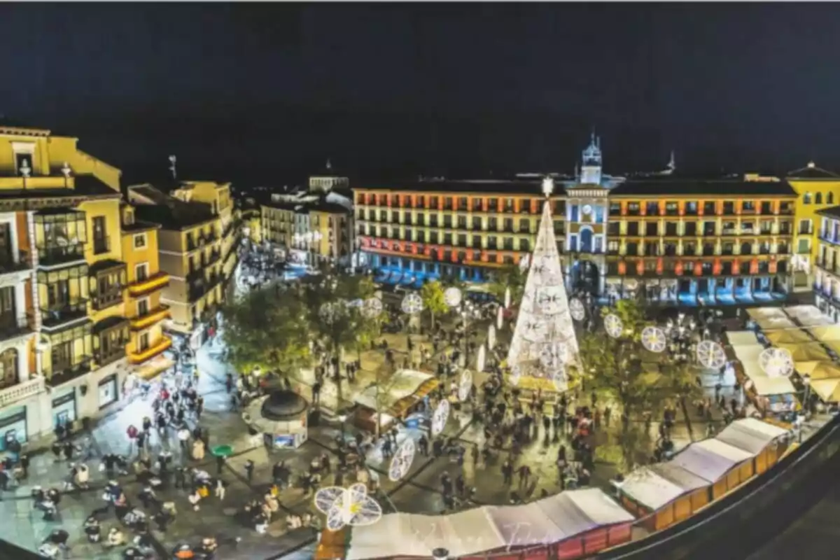 Vista nocturna de una plaza iluminada con un árbol de Navidad y edificios alrededor.