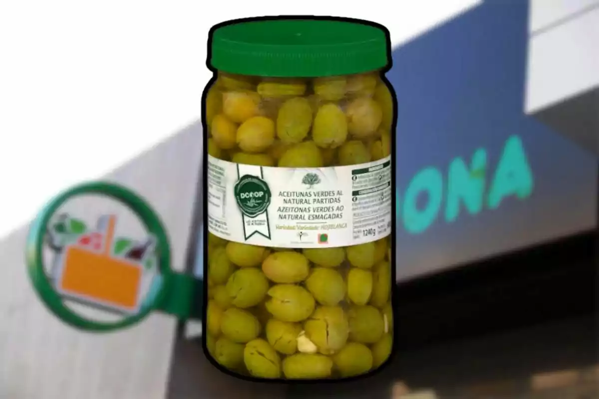 Un frasco de aceitunas verdes al natural partidas con tapa verde y etiqueta blanca frente a un fondo borroso de un supermercado. Un frasco de aceitunas verdes al natural partidas con tapa verde y etiqueta blanca frente a un fondo borroso de un supermercado.