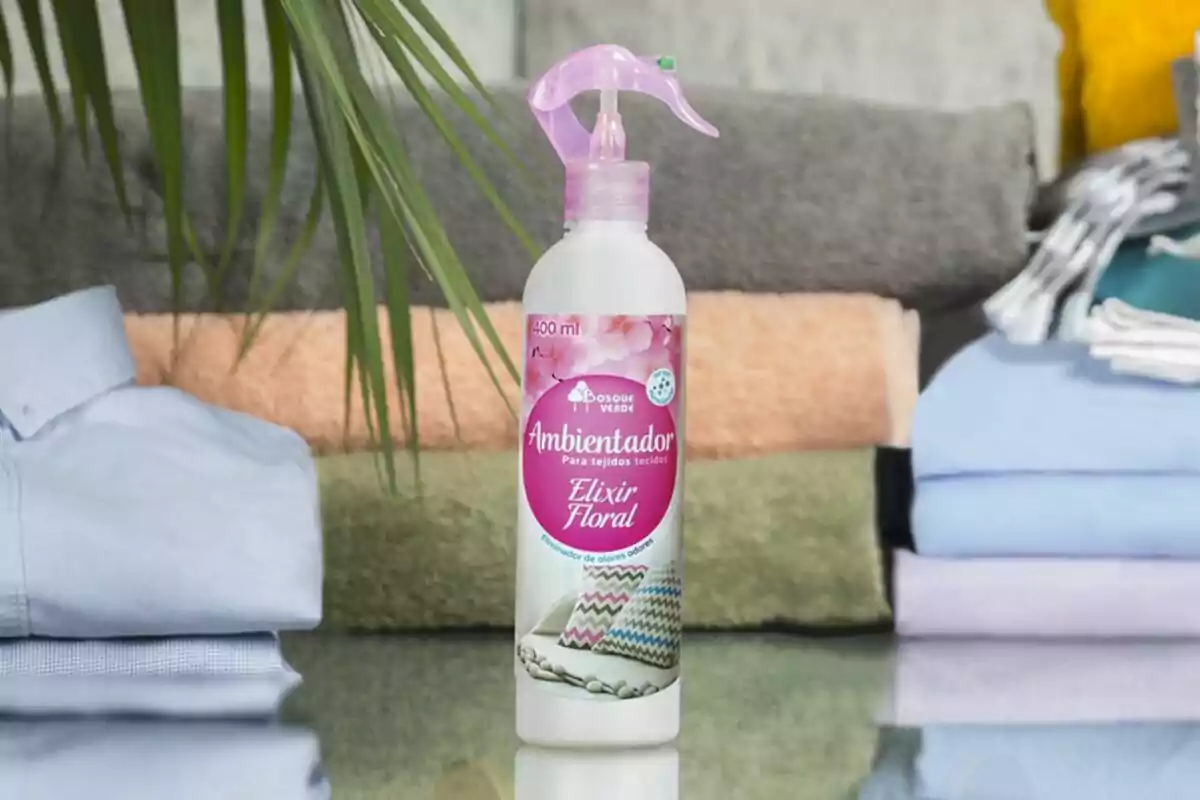 Ambientador para tejidos con fragancia Elixir Floral de 400 ml, colocado sobre una superficie de vidrio con toallas y ropa doblada en el fondo. Ambientador para tejidos con fragancia Elixir Floral de 400 ml, colocado sobre una superficie de vidrio con toallas y ropa doblada en el fondo.