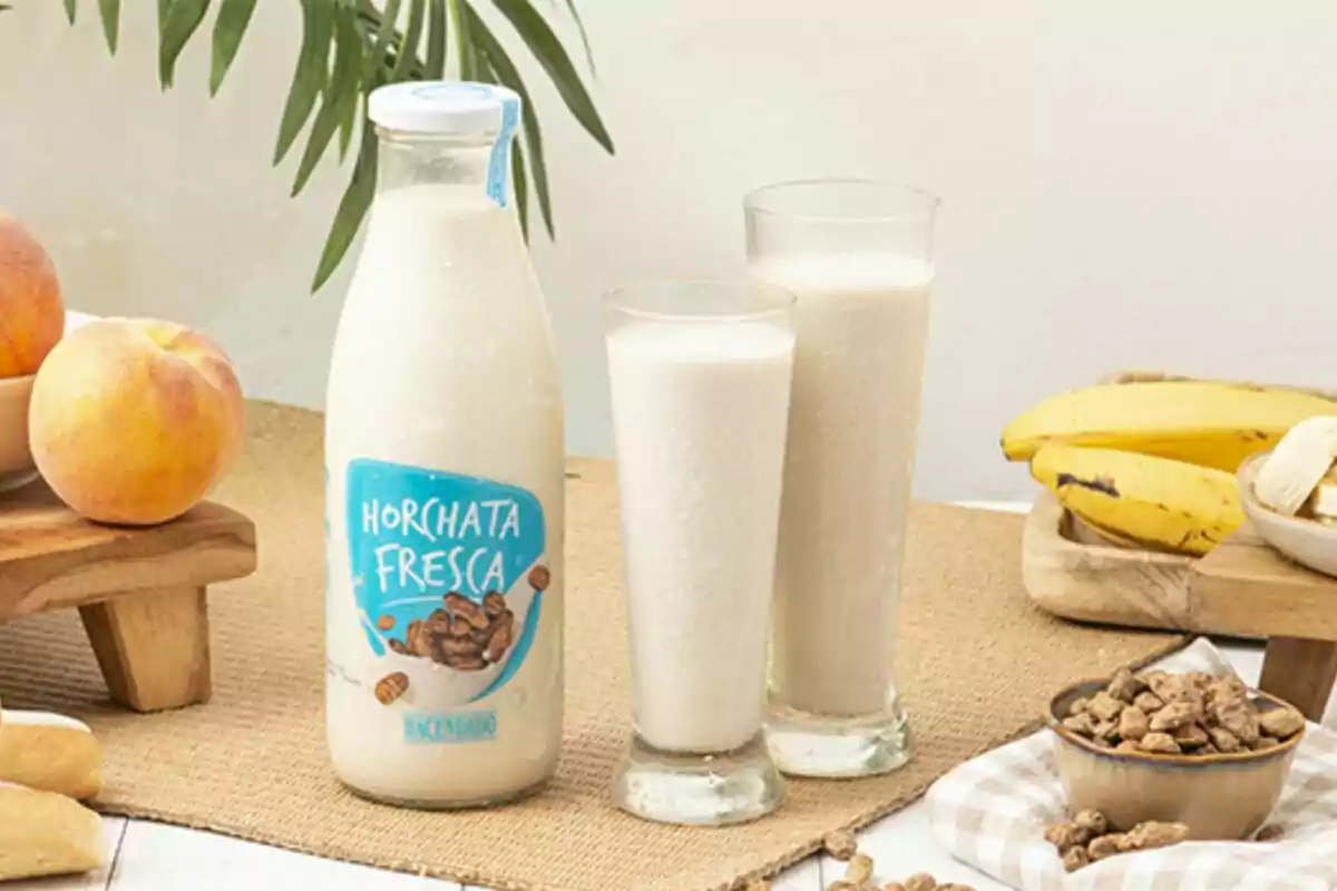 Una botella de horchata fresca junto a dos vasos llenos de la bebida rodeados de frutas como melocotones y plátanos sobre una mesa. Una botella de horchata fresca junto a dos vasos llenos de la bebida rodeados de frutas como melocotones y plátanos sobre una mesa.