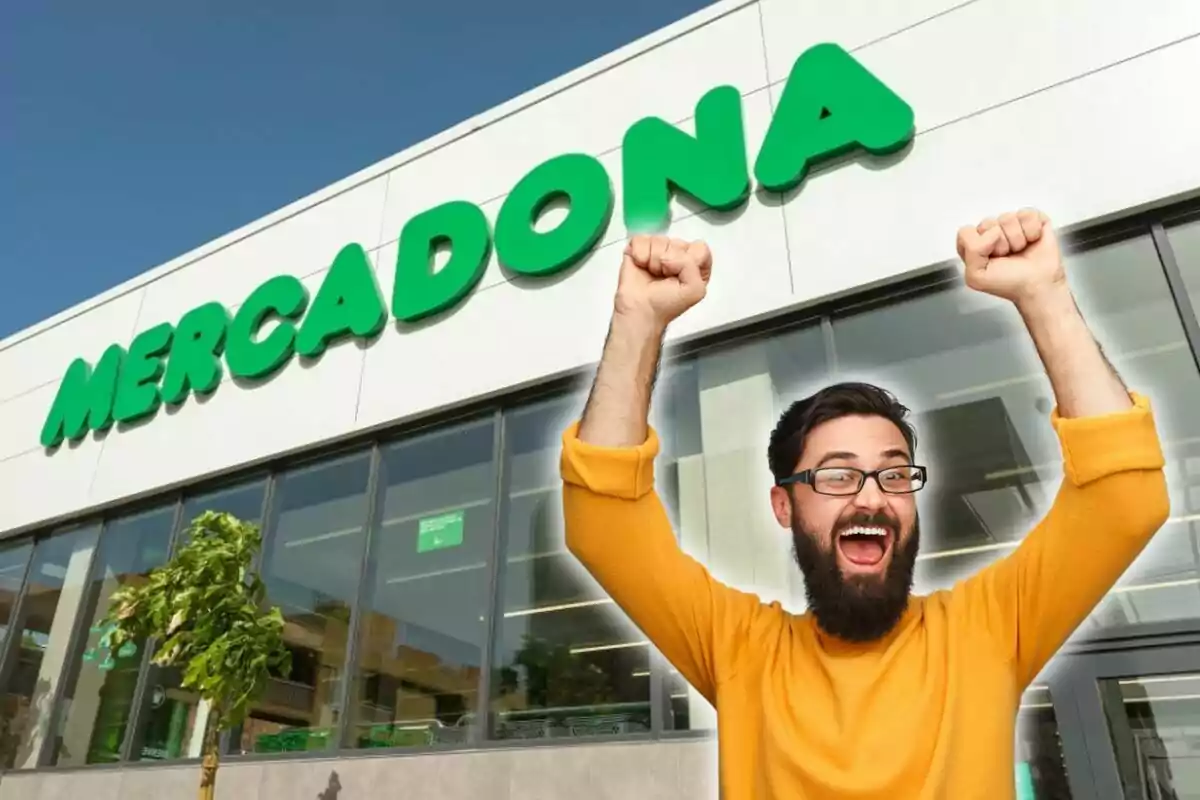 Un hombre con barba y gafas, vestido con un suéter amarillo, celebra con los brazos en alto frente a una tienda de Mercadona.