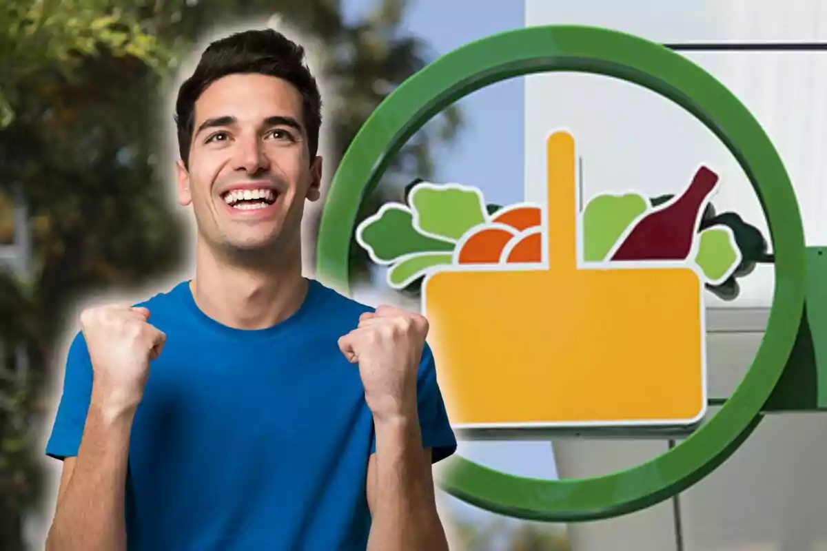 Hombre joven sonriente con camiseta azul celebrando frente a un letrero de supermercado con una canasta de compras llena de alimentos