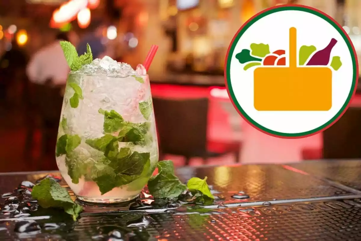 Un cóctel con hojas de menta y hielo en un vaso sobre una mesa mojada en un ambiente de bar con un logotipo de cesta de alimentos al lado. Un cóctel con hojas de menta y hielo en un vaso sobre una mesa mojada en un ambiente de bar con un logotipo de cesta de alimentos al lado.