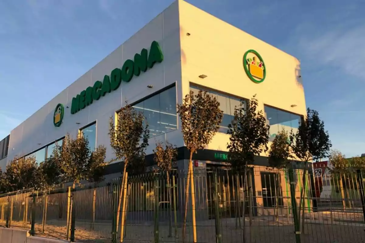 Edificio de supermercado Mercadona con logotipo verde y amarillo en la fachada, rodeado de árboles y una cerca. Edificio de supermercado Mercadona con logotipo verde y amarillo en la fachada, rodeado de árboles y una cerca.