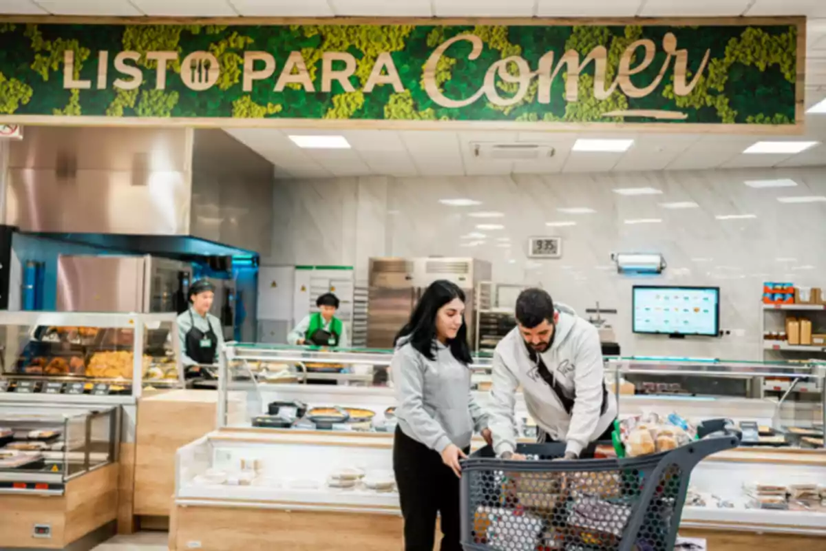 Una pareja con un carrito de compras en una sección de comida preparada de un supermercado con un letrero que dice Una pareja con un carrito de compras en una sección de comida preparada de un supermercado con un letrero que dice