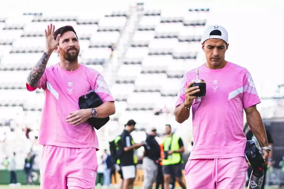 Dos personas con ropa deportiva rosa caminan en un estadio, una de ellas saluda mientras la otra sostiene un mate. Dos personas con ropa deportiva rosa caminan en un estadio, una de ellas saluda mientras la otra sostiene un mate.