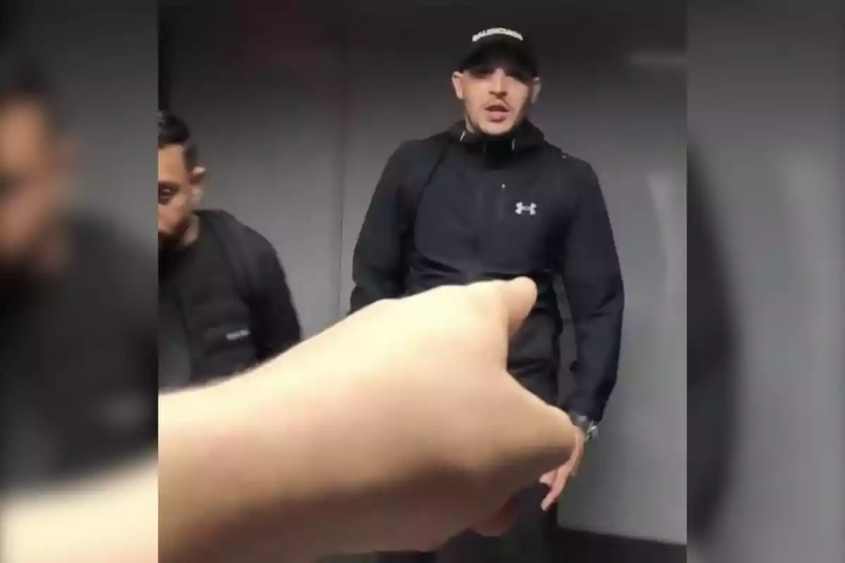 Una mano en primer plano señala a un hombre con gorra y ropa deportiva negra que está de pie junto a otra persona en un entorno interior.
