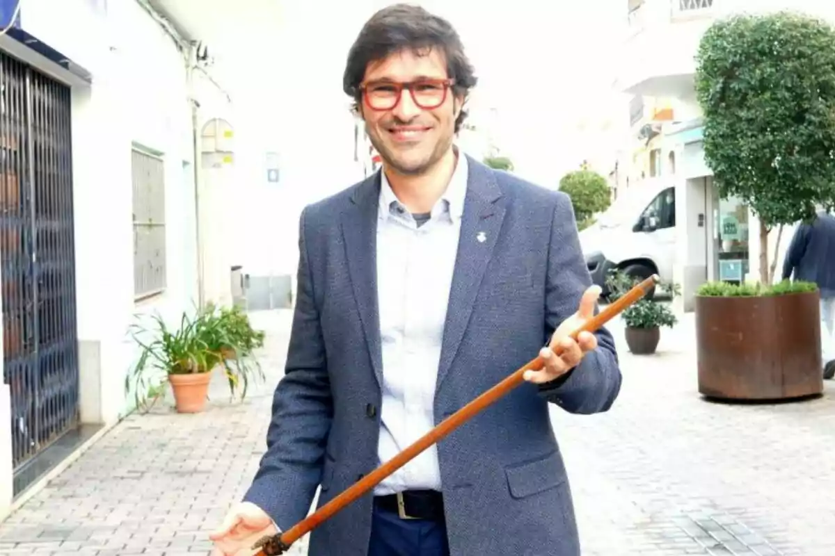 Un hombre con gafas rojas y traje azul sostiene un bastón en una calle peatonal. Un hombre con gafas rojas y traje azul sostiene un bastón en una calle peatonal.