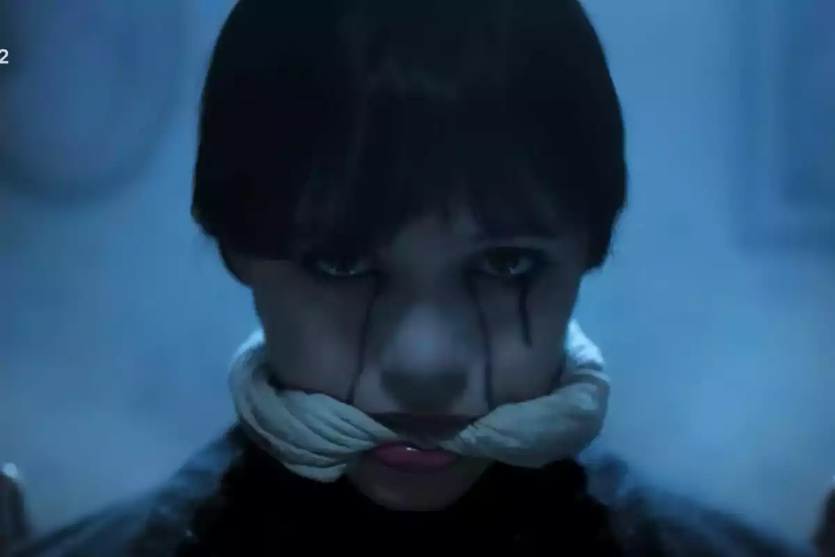 Jenna Ortega con expresión seria y maquillaje oscuro en los ojos, con una tela blanca atada alrededor de la boca en un ambiente de luz azulada en la serie Miércoles