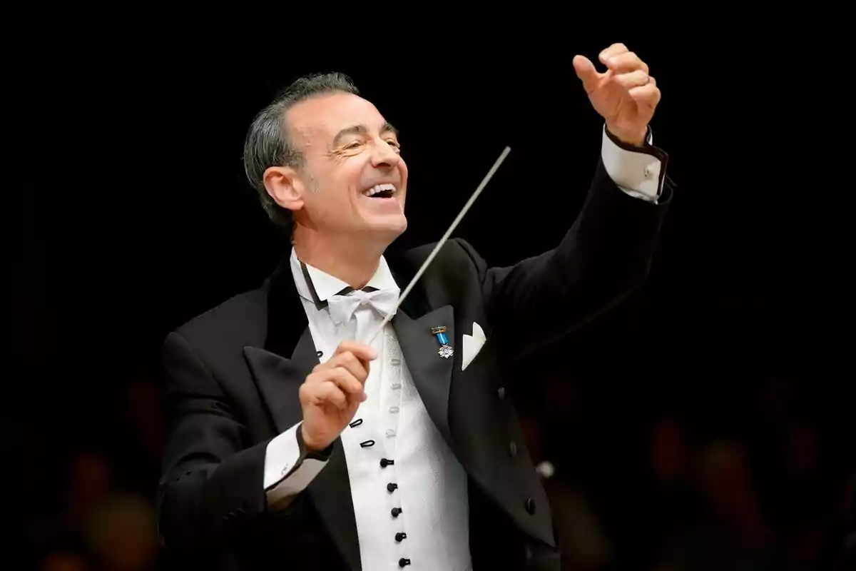 Fotografía de Miguel Ángel Gómez-Martínez como director de orquesta sonriente dirigiendo con una batuta en la mano.