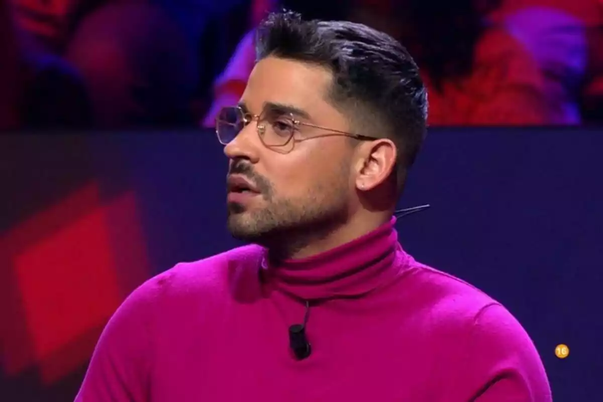 Miguel Frigenti con gafas y barba lleva un suéter fucsia y está en un entorno iluminado con luces de colores en Gran Hermano.