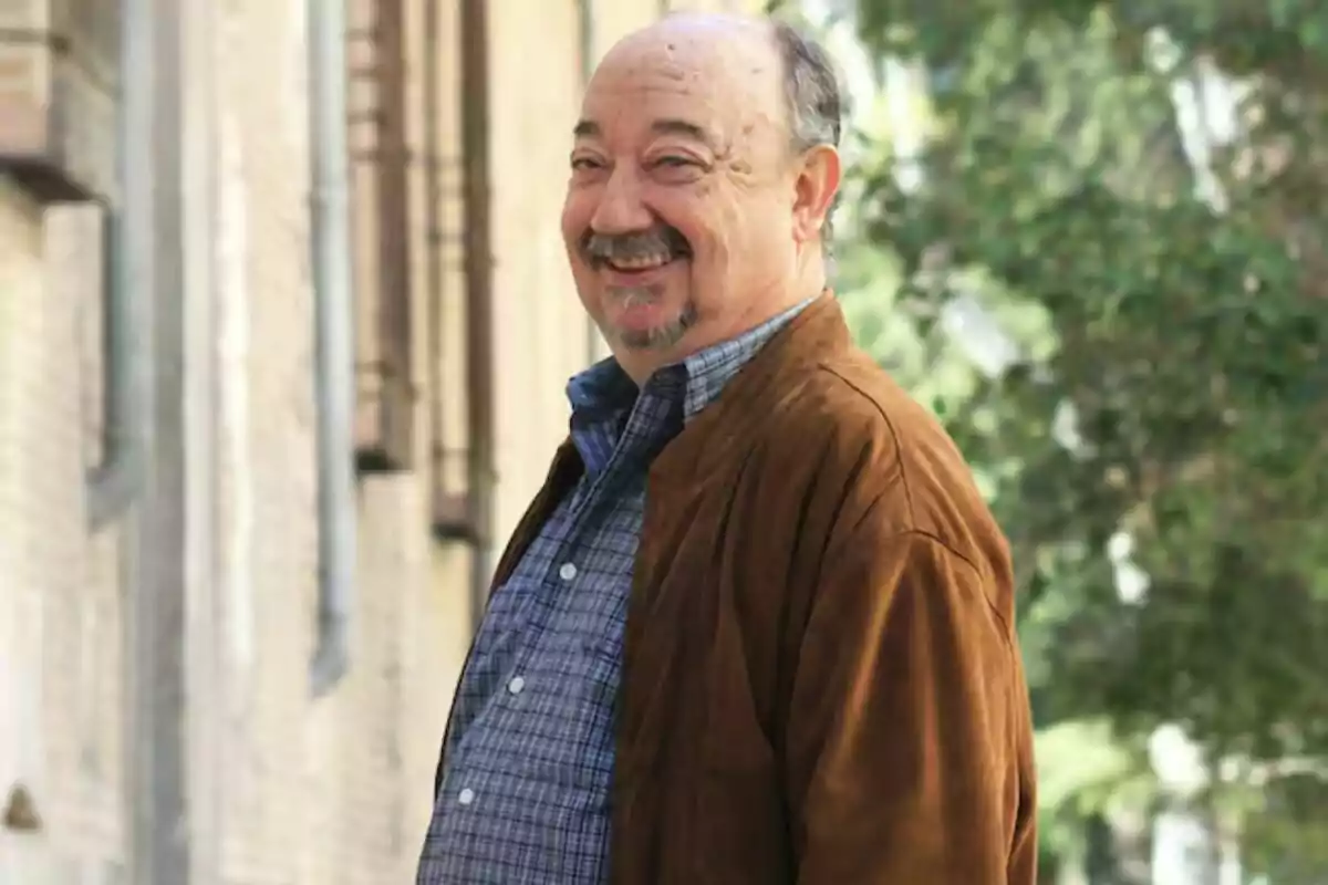 Miguel Vigil sonriendo con una chaqueta marrón y camisa a cuadros, de pie en una calle con árboles y edificios de fondo.