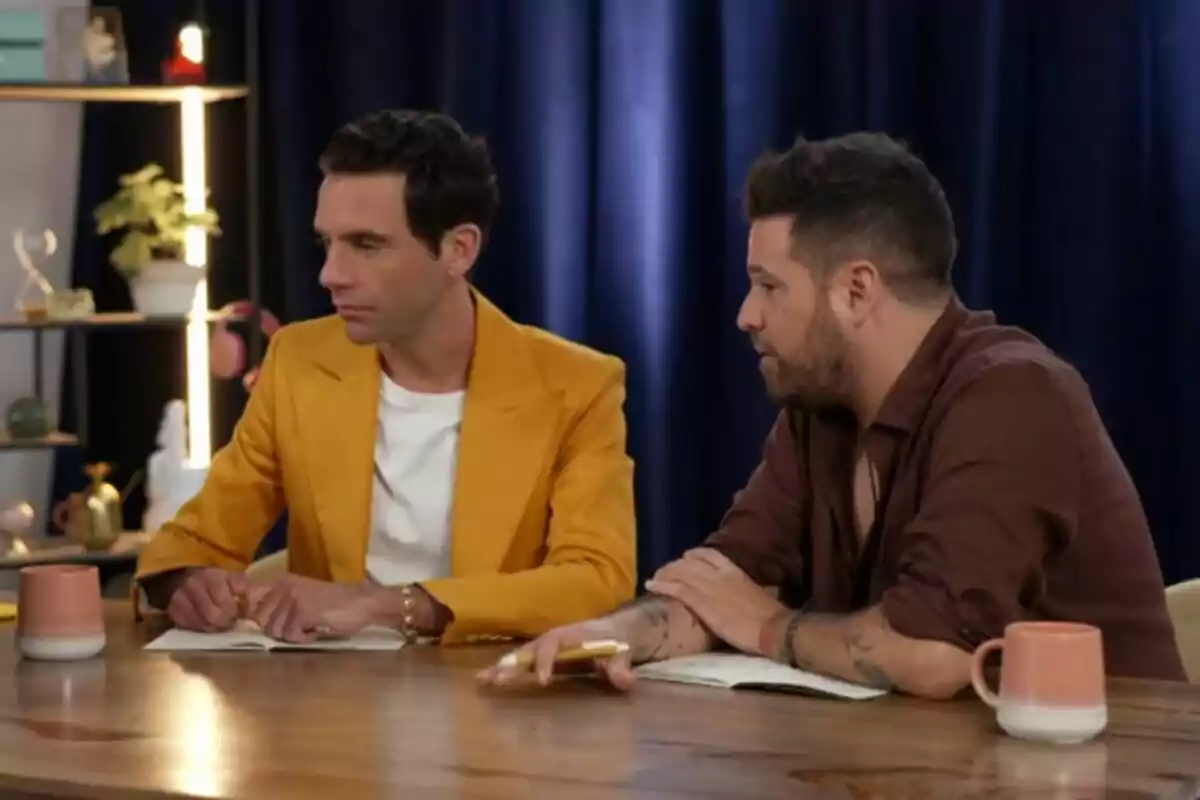 Mika y Pablo López sentadas en una mesa con tazas y cuadernos, una lleva un saco amarillo y la otra una camisa marrón, con un fondo de cortinas azules y estanterías en El Piano.