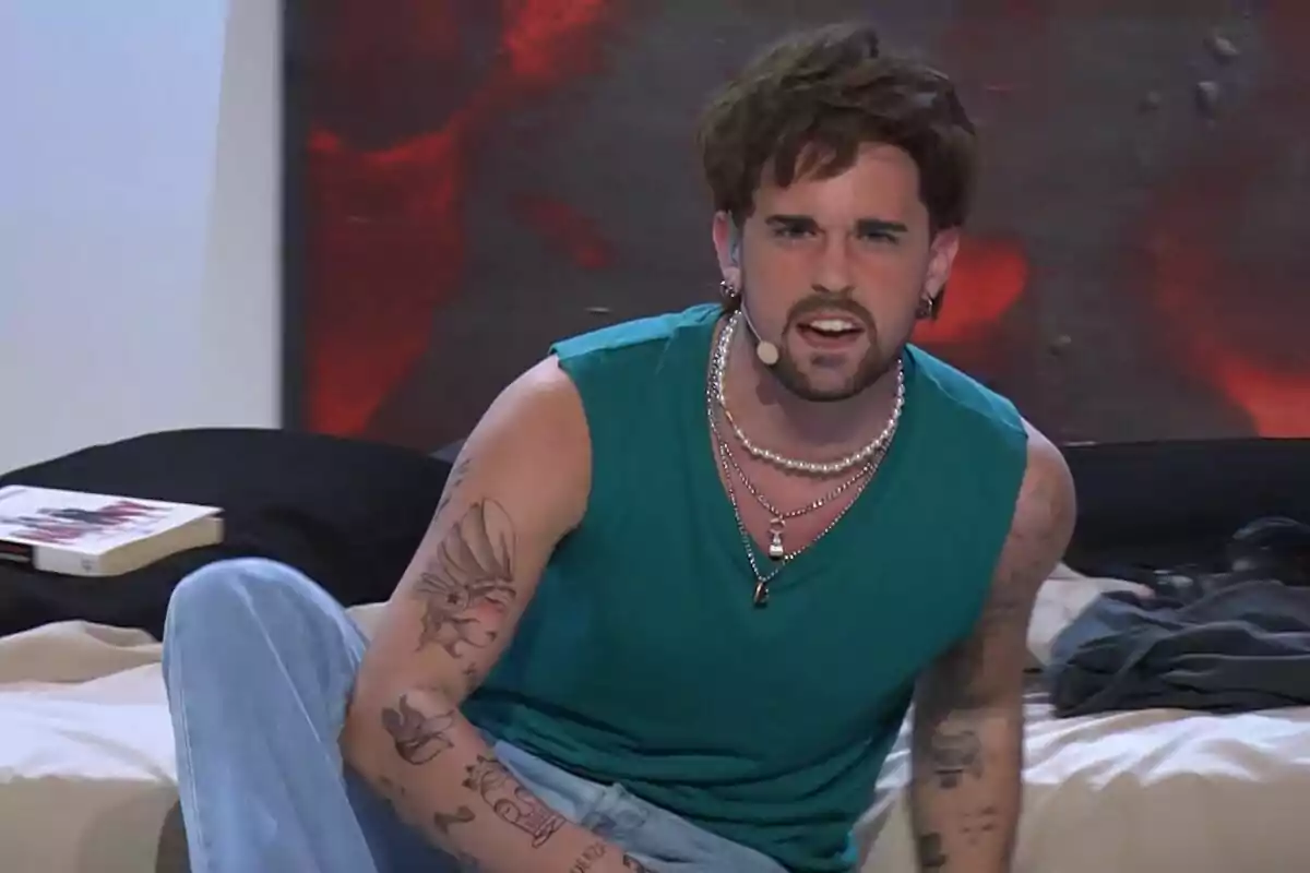 Mikel Herzog JR con barba y tatuajes en los brazos está sentado en una cama, lleva una camiseta sin mangas verde y un micrófono de diadema en Tu Cara Me Suena. Mikel Herzog JR con barba y tatuajes en los brazos está sentado en una cama, lleva una camiseta sin mangas verde y un micrófono de diadema en Tu Cara Me Suena.