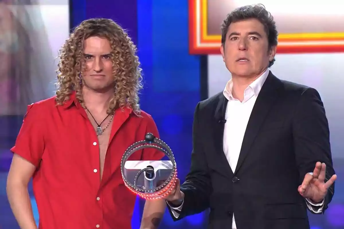 Mikel Herzog y Manel Fuentes de pie en un escenario de Tu Cara Me Suena, una con camisa roja y cabello rizado, la otra con traje oscuro y camisa blanca, sosteniendo un objeto circular.