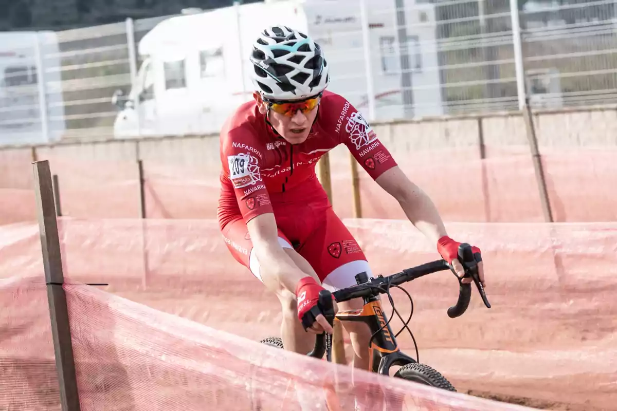 Ciclista con uniforme rojo y casco blanco compitiendo en una carrera de ciclismo en un circuito delimitado por mallas rosas Ciclista con uniforme rojo y casco blanco compitiendo en una carrera de ciclismo en un circuito delimitado por mallas rosas