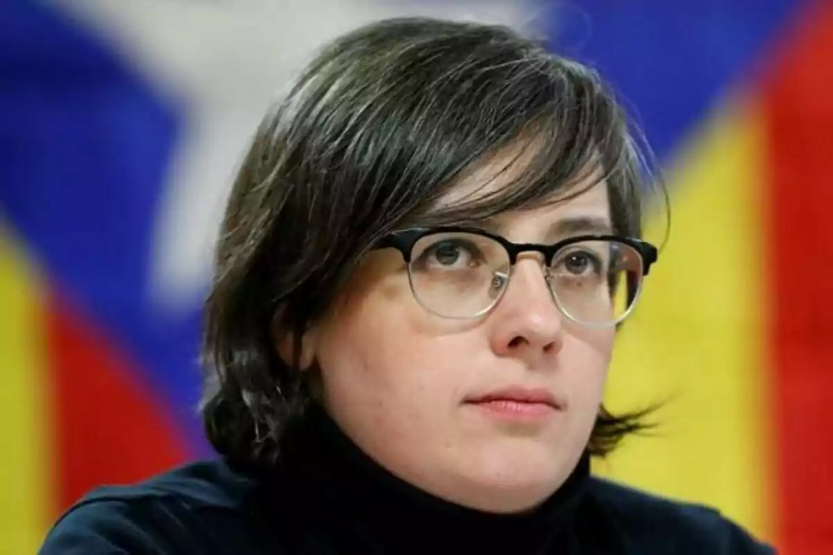 Una persona con gafas y cabello oscuro frente a un fondo con colores azul, amarillo y rojo.