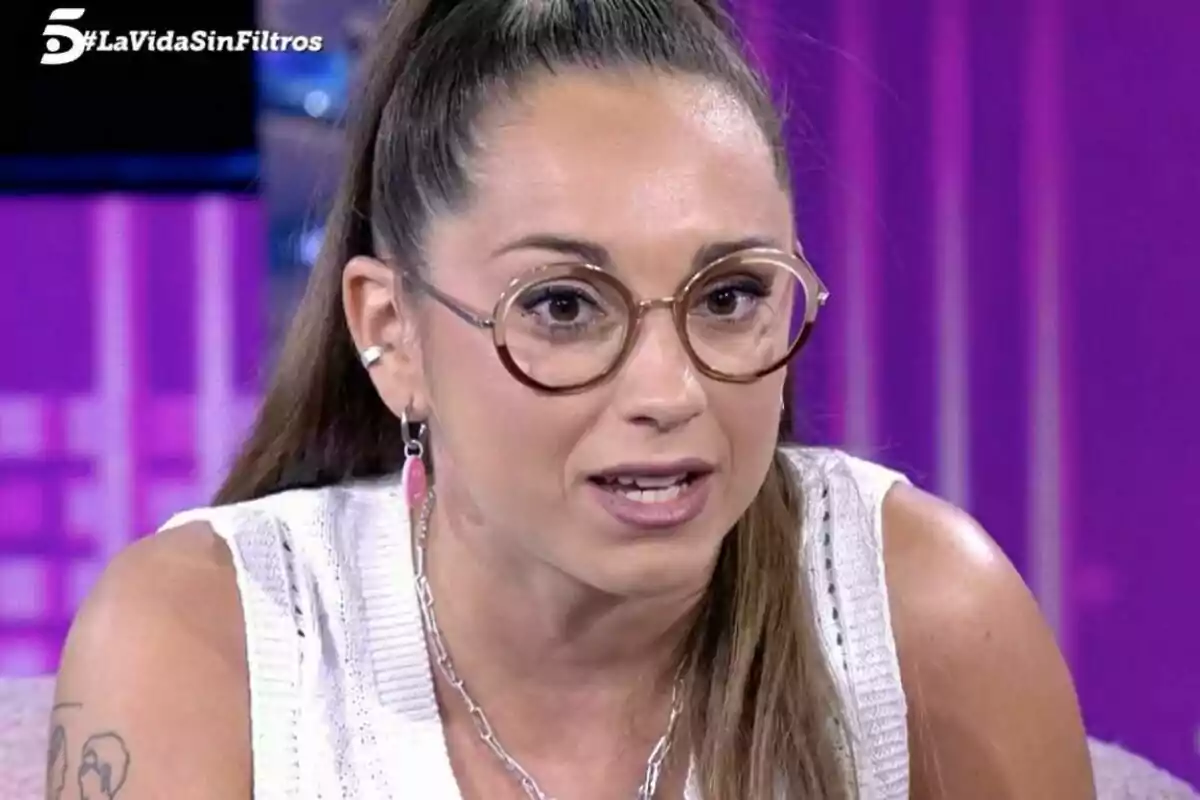 Captura de Miriam Corregüela con gafas redondas y cabello recogido en una coleta alta, lleva un suéter blanco sin mangas y está sentada en el plató de La Vida Sin Filtros con fondo morado.