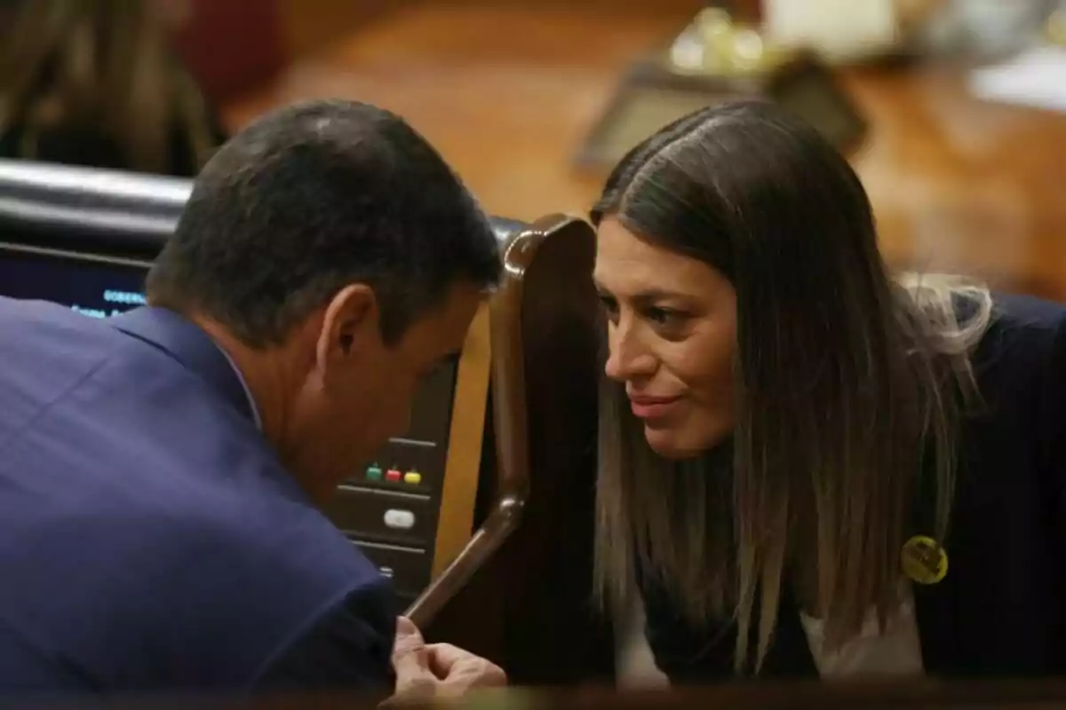 Dos personas conversan en un entorno formal, inclinadas hacia adelante para hablar en privado