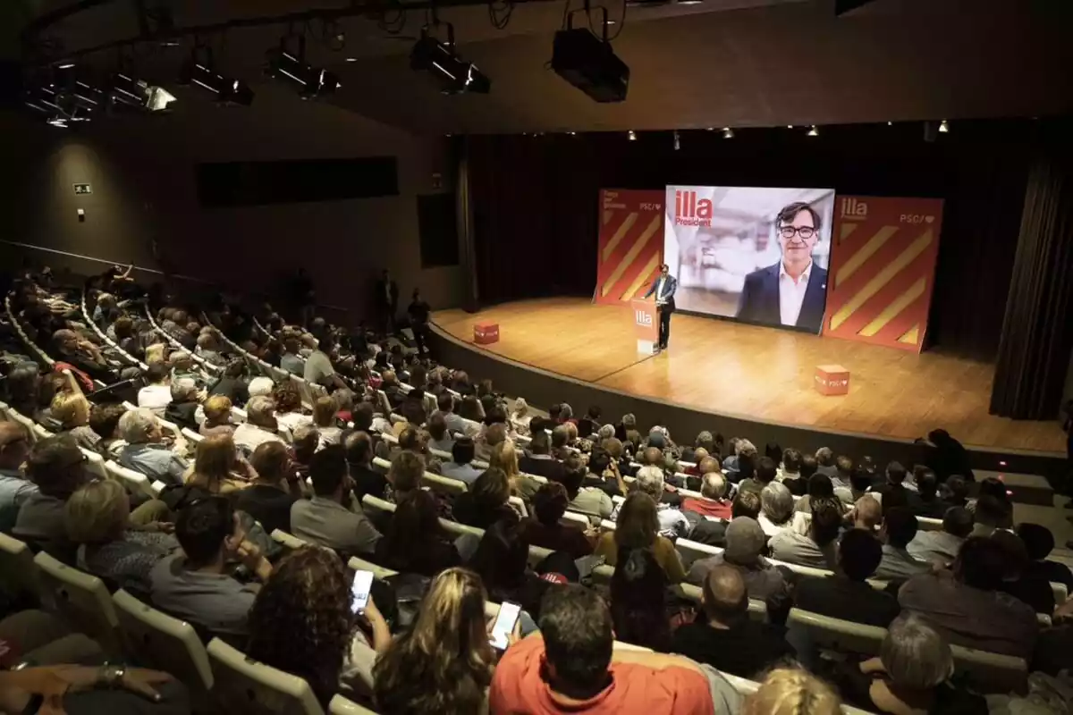 Plano general de una sala con decenas de personas sentadas en las gradas y Salvador Illa, solo delante de un atril en el escenario situado a la parte derecha de la imagen