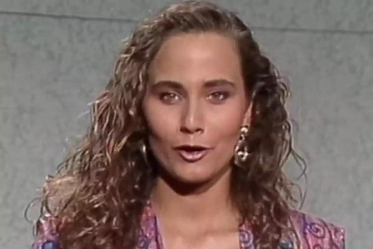 Mujer con cabello rizado y largo, usando aretes grandes y una chaqueta colorida, hablando frente a una pared gris. Mujer con cabello rizado y largo, usando aretes grandes y una chaqueta colorida, hablando frente a una pared gris.