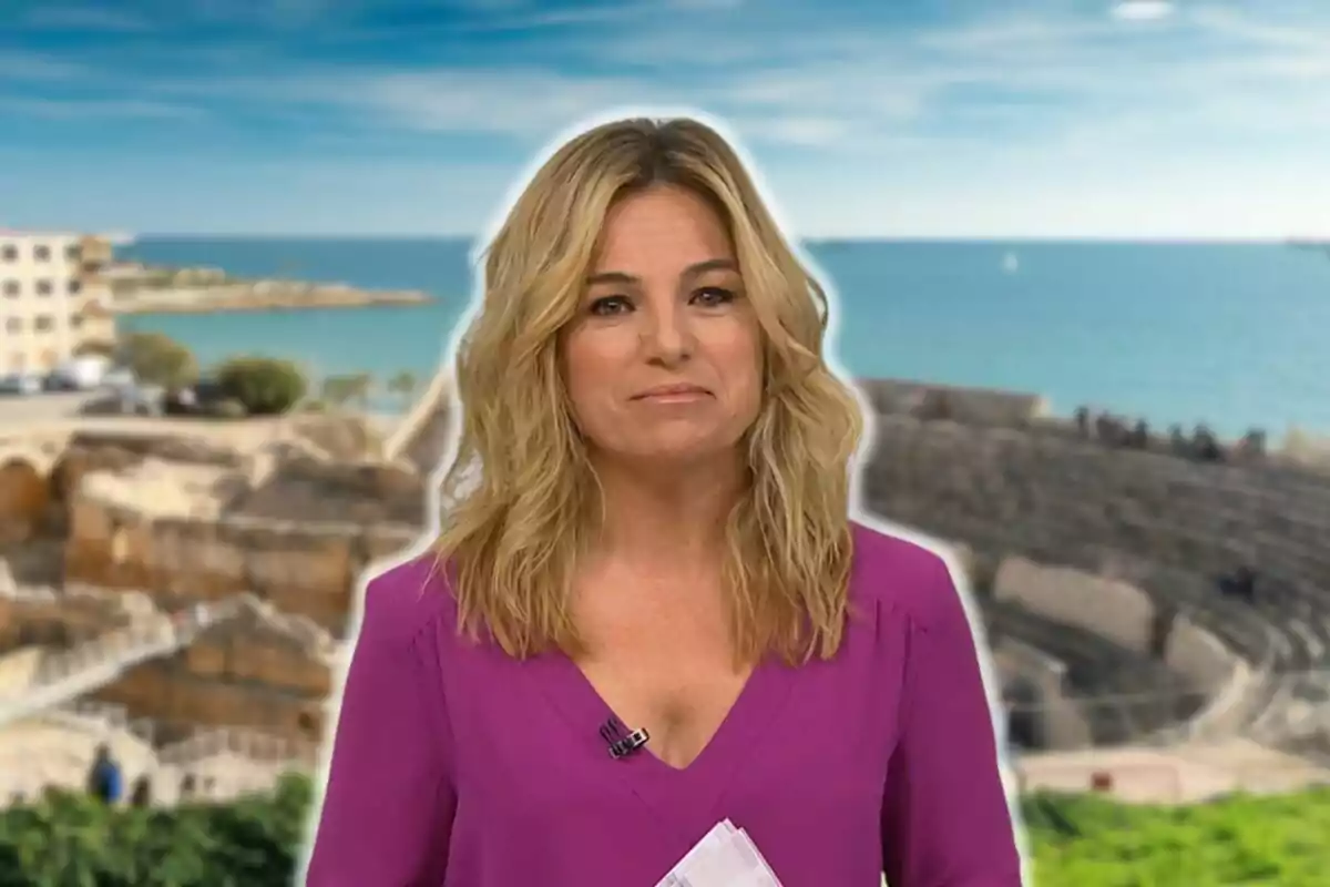 Núria Solé rubia con una blusa morada está de pie frente a un fondo que muestra un paisaje costero con ruinas antiguas y el mar al fondo.
