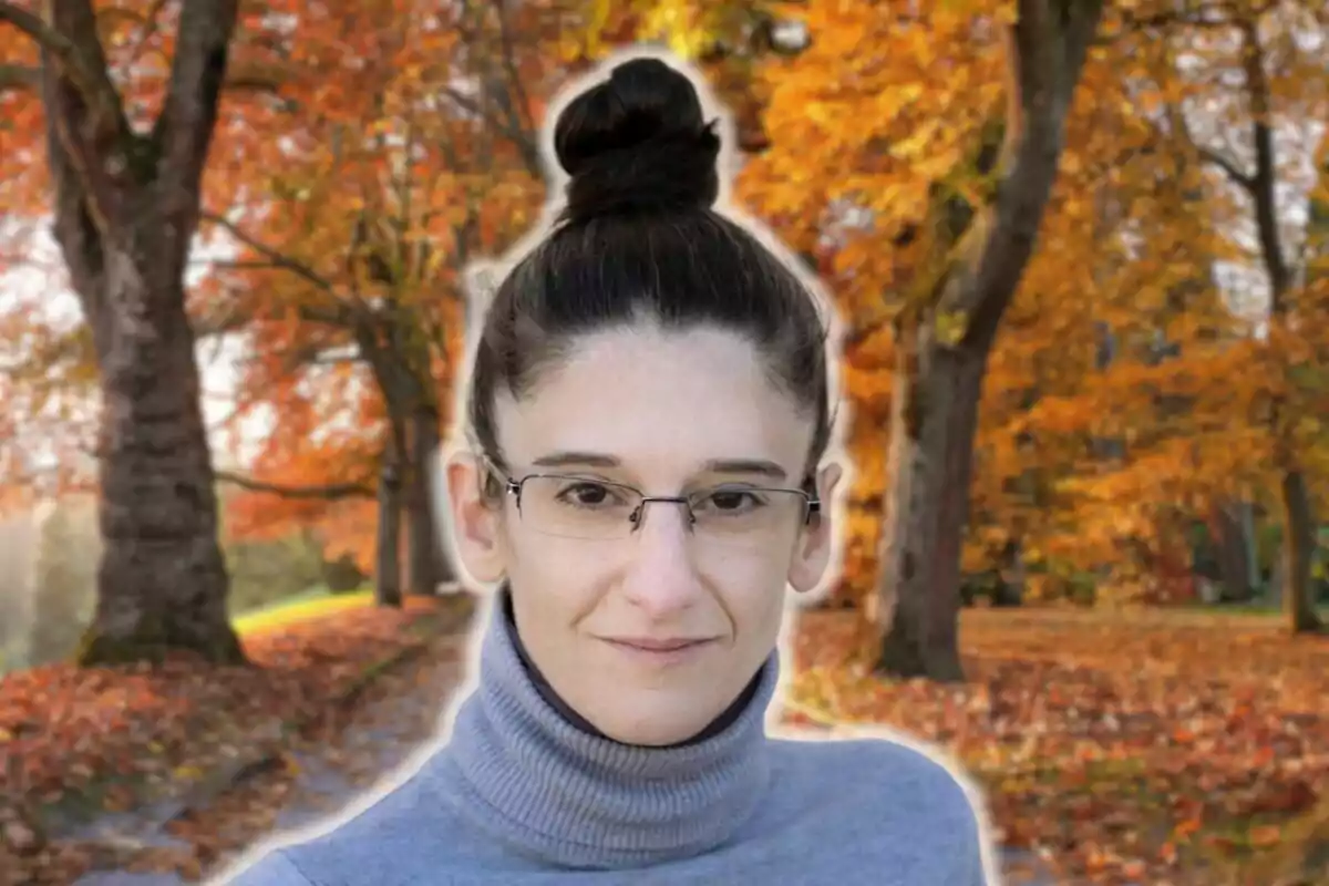 Maria Nicolau con gafas y cabello recogido en un moño posa frente a un paisaje otoñal con árboles de hojas naranjas.