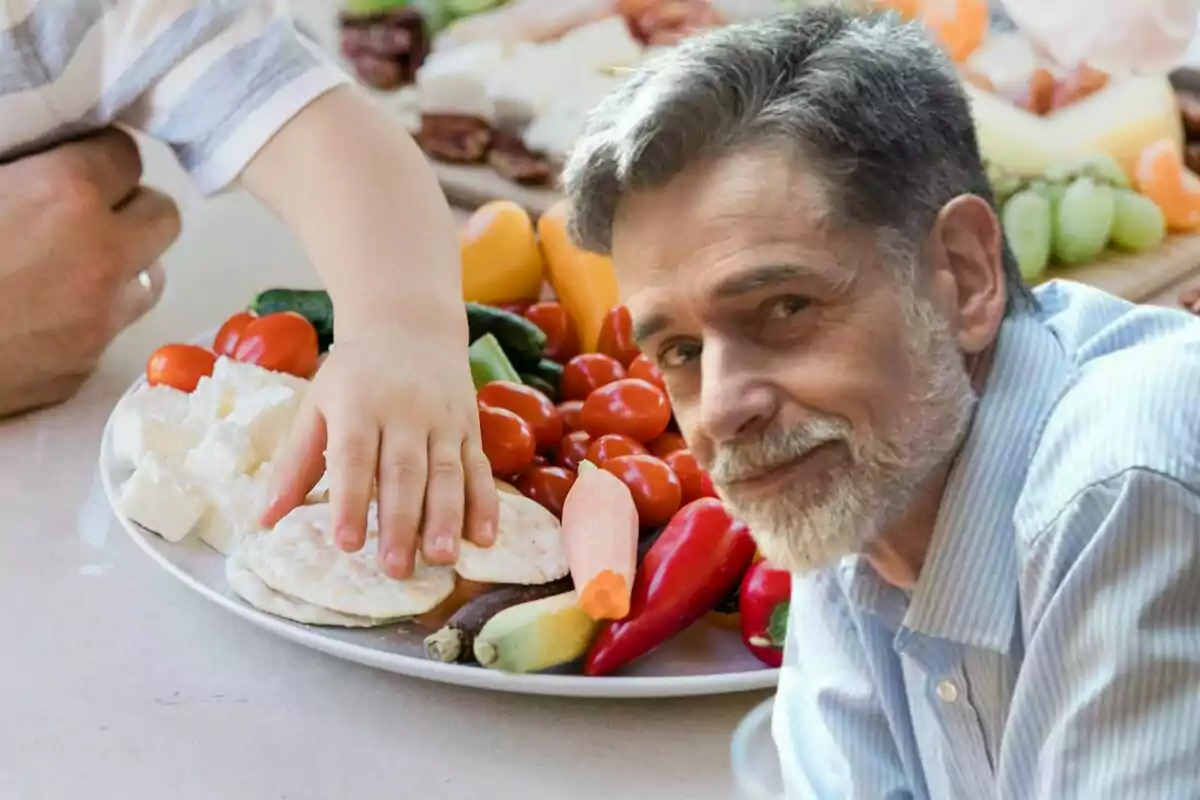 Un niño pequeño toca un plato lleno de verduras y queso mientras un hombre mayor sonríe al frente.