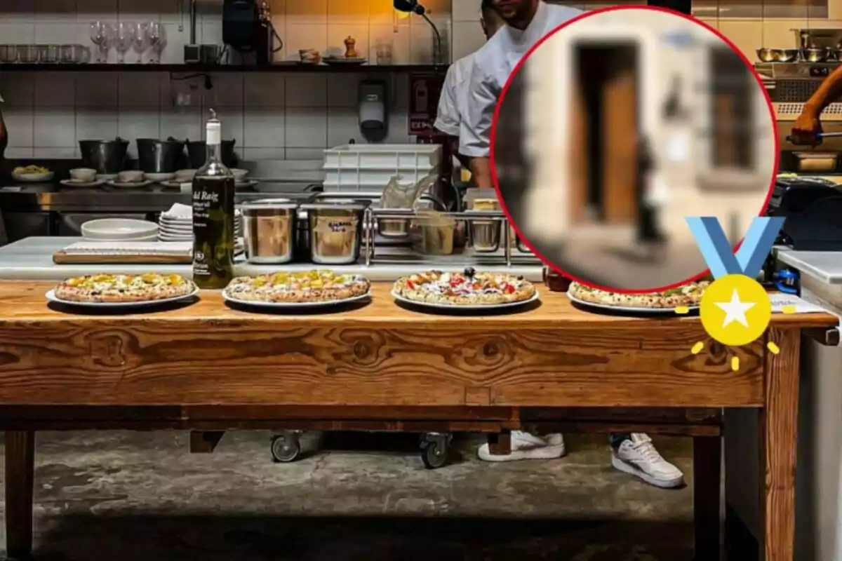 Una mesa de madera en una cocina con varias pizzas listas para servir y utensilios de cocina al fondo.