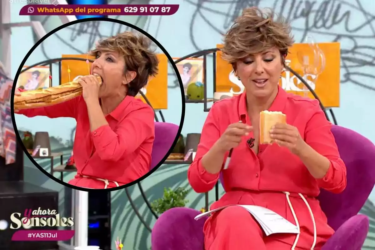 Una mujer con camisa roja está sentada en un sillón morado, sosteniendo y comiendo un bocadillo grande en un programa de televisión.