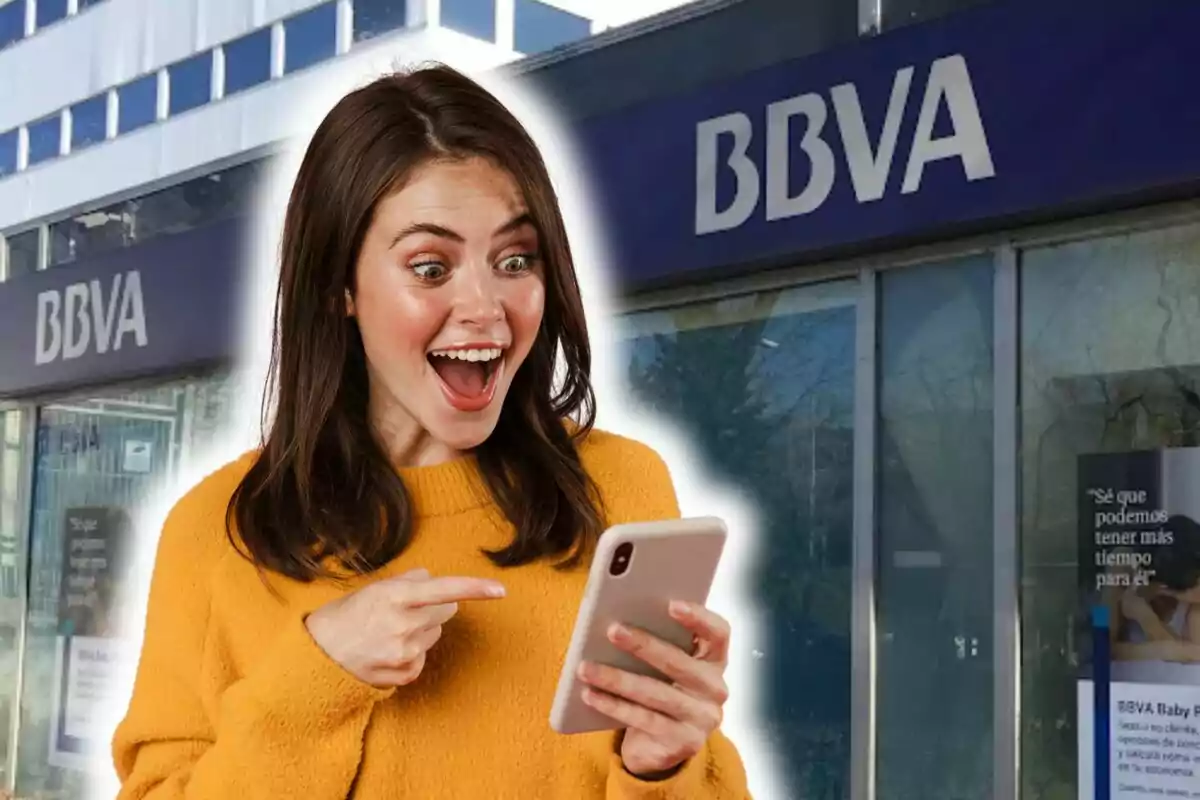 Una mujer con suéter amarillo mira emocionada su teléfono frente a una sucursal de BBVA.