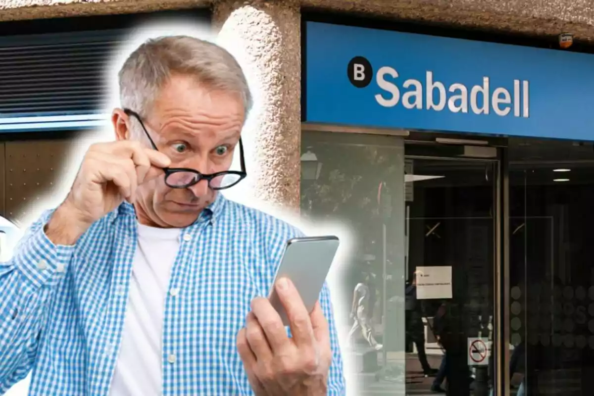 Un hombre mayor con una camisa a cuadros azules y gafas mira sorprendido su teléfono móvil frente a una sucursal del banco Sabadell.