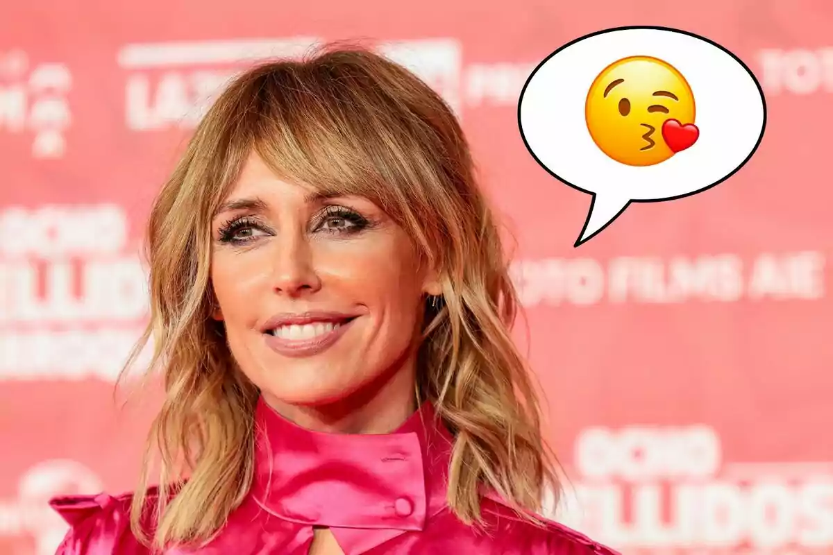 Emma García sonriente con cabello rubio y ondulado, vestida con una blusa rosa brillante, con un fondo rojo y un emoji de beso en un globo de diálogo.