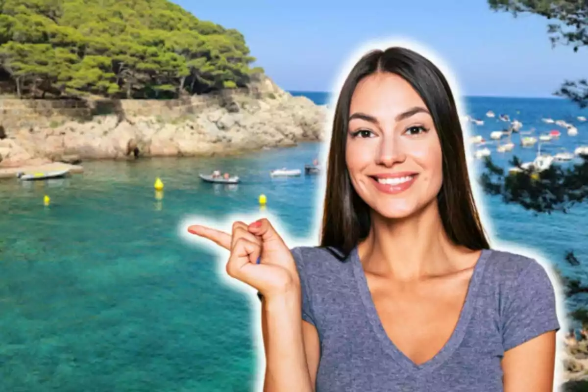 Una mujer sonriente con cabello largo y oscuro, vestida con una camiseta gris, señala con su dedo índice hacia un paisaje costero con agua cristalina, pequeñas embarcaciones y una colina cubierta de árboles verdes ubicado en Cala Sa Tuna (Costa Brava, Gerona).