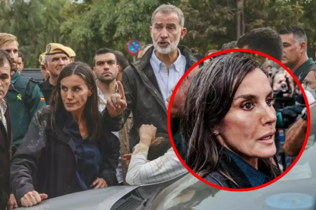 Un grupo de personas, algunas con uniforme, se encuentra en un entorno al aire libre; Letizia Ortiz en primer plano parece estar hablando o señalando algo, mientras que Felipe de Borbón detrás de ella también parece estar hablando.