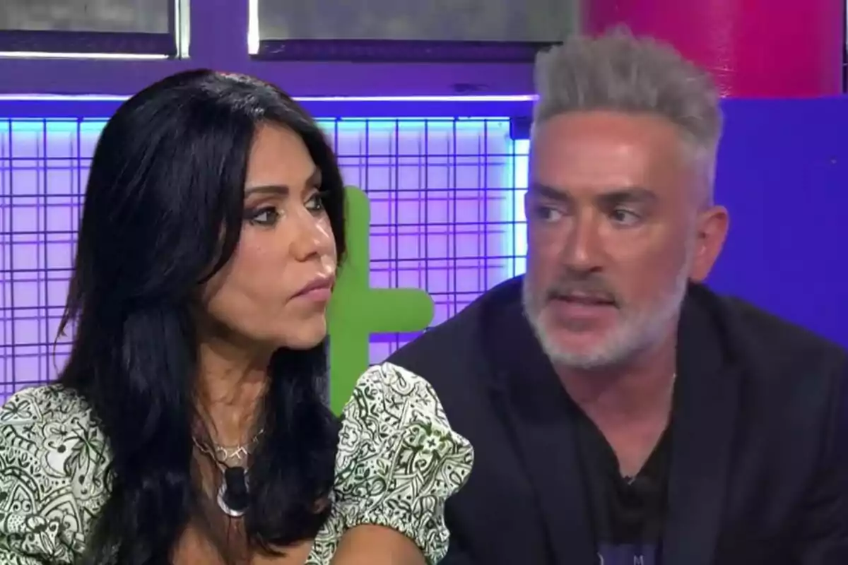 Maite Galdeano y Kiko Hernández conversando en un set de televisión con fondo colorido.