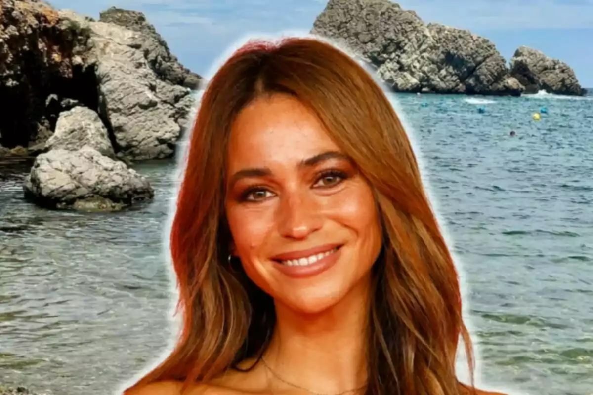 María Verdoy sonriente con cabello castaño claro y ondulado está frente a un fondo de mar y rocas.