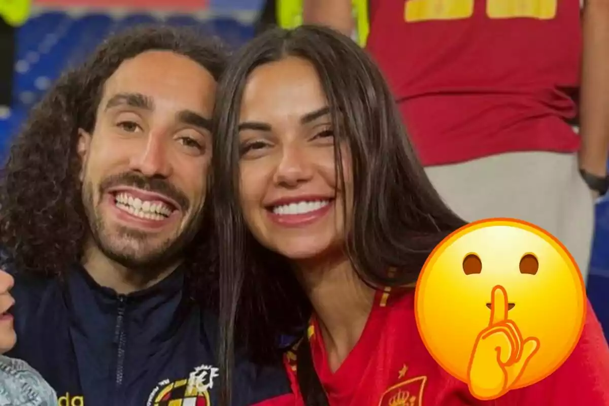 Marc Cucurella y Claudia Rodríguez sonrientes posan para una foto, Marc lleva una chaqueta con el escudo de la Federación Española de Fútbol y Claudia la camiseta, con un emoji de "silencio" superpuesto en la esquina inferior derecha.