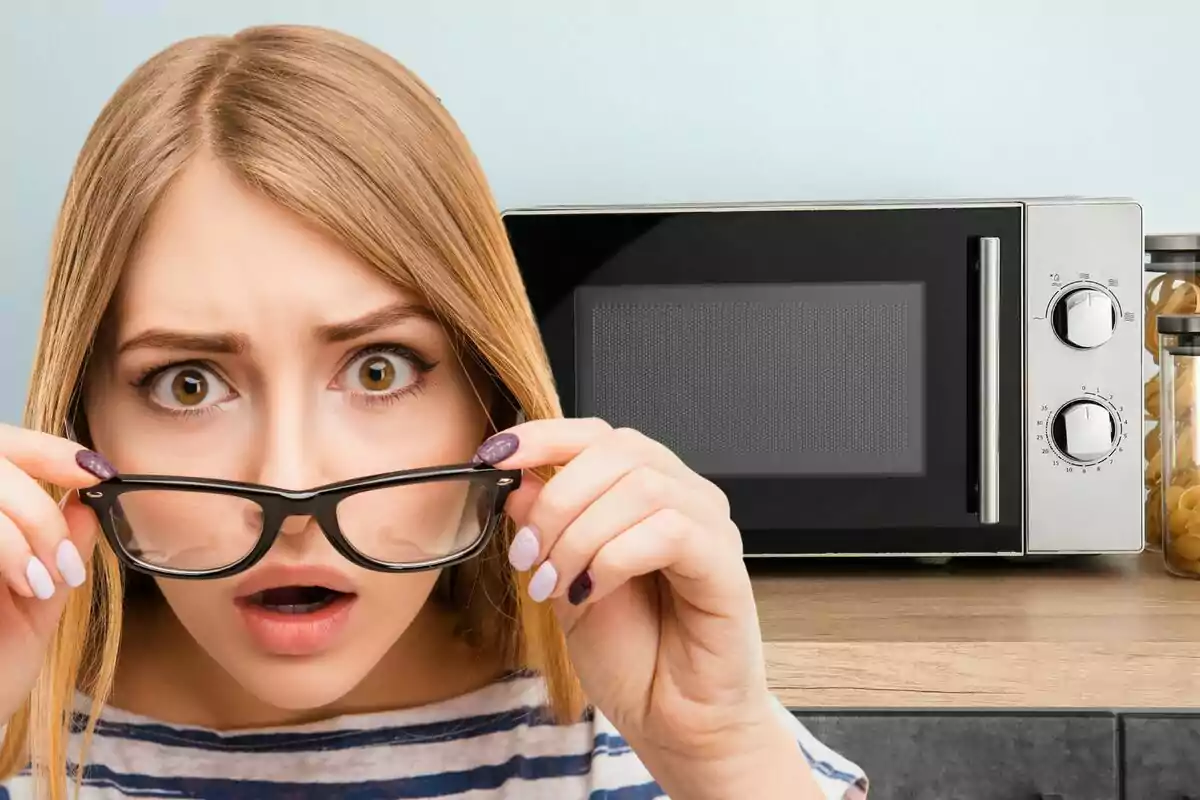 Mujer sorprendida sosteniendo sus gafas frente a un microondas en una cocina.