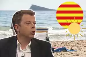 Francesc Mauri con traje oscuro hablando frente a una playa con una silla y una toalla en la arena, una bandera de Cataluña y un sol.