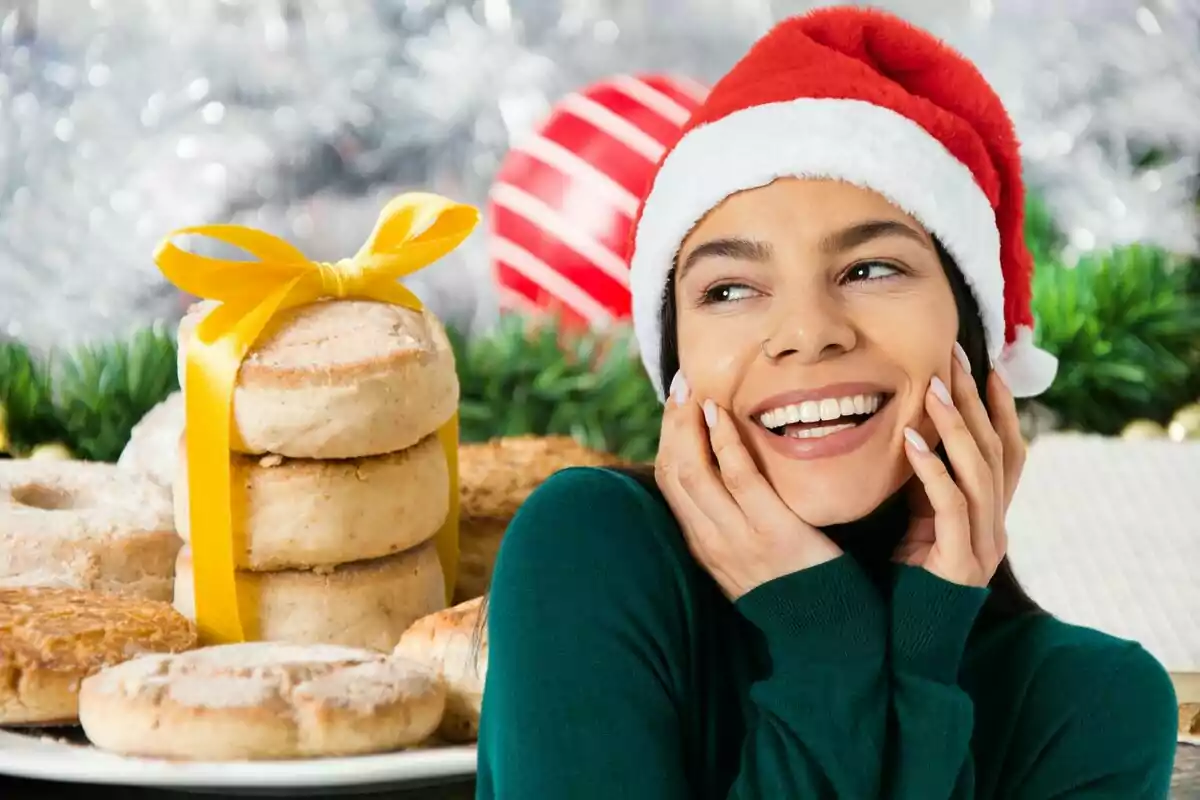 Una persona sonriente con un gorro de Navidad junto a una pila de galletas decoradas con un lazo amarillo, con un fondo festivo.