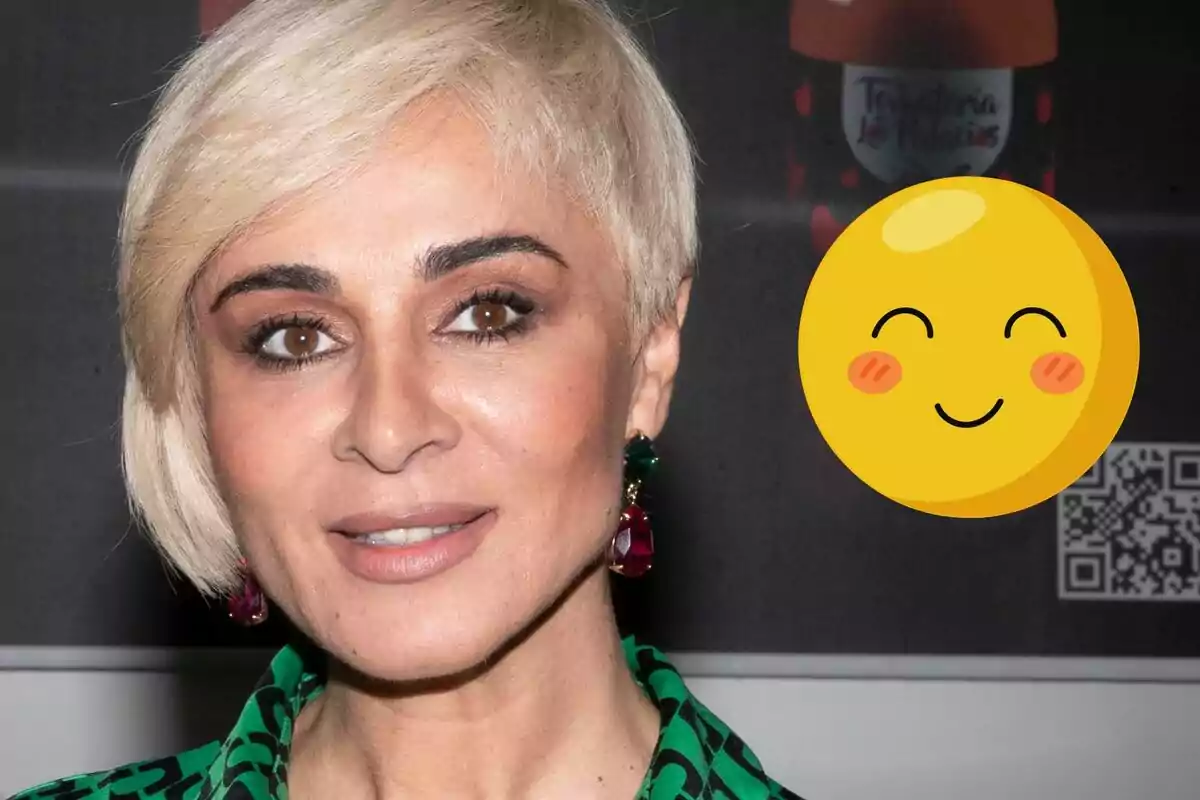 Ana María Aldón con cabello rubio corto y aretes coloridos sonríe frente a un fondo oscuro con un emoji sonriente superpuesto.