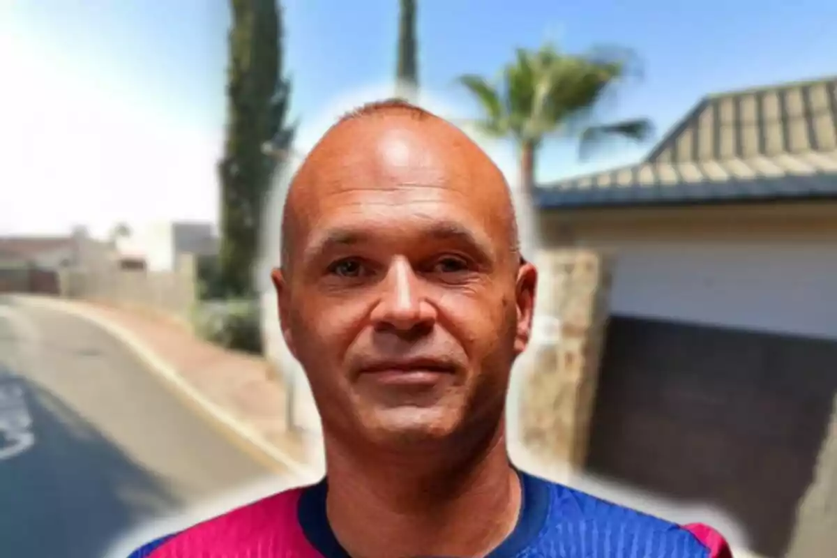 Andrés Iniesta con camiseta azul y roja posando frente a una calle con árboles y casas al fondo.