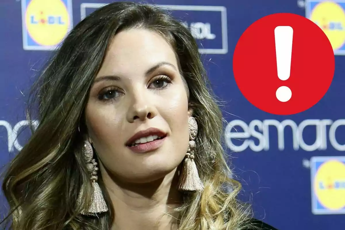 Jessica Bueno con pendientes grandes frente a un fondo azul con logotipos y un círculo rojo con un signo de exclamación blanco.