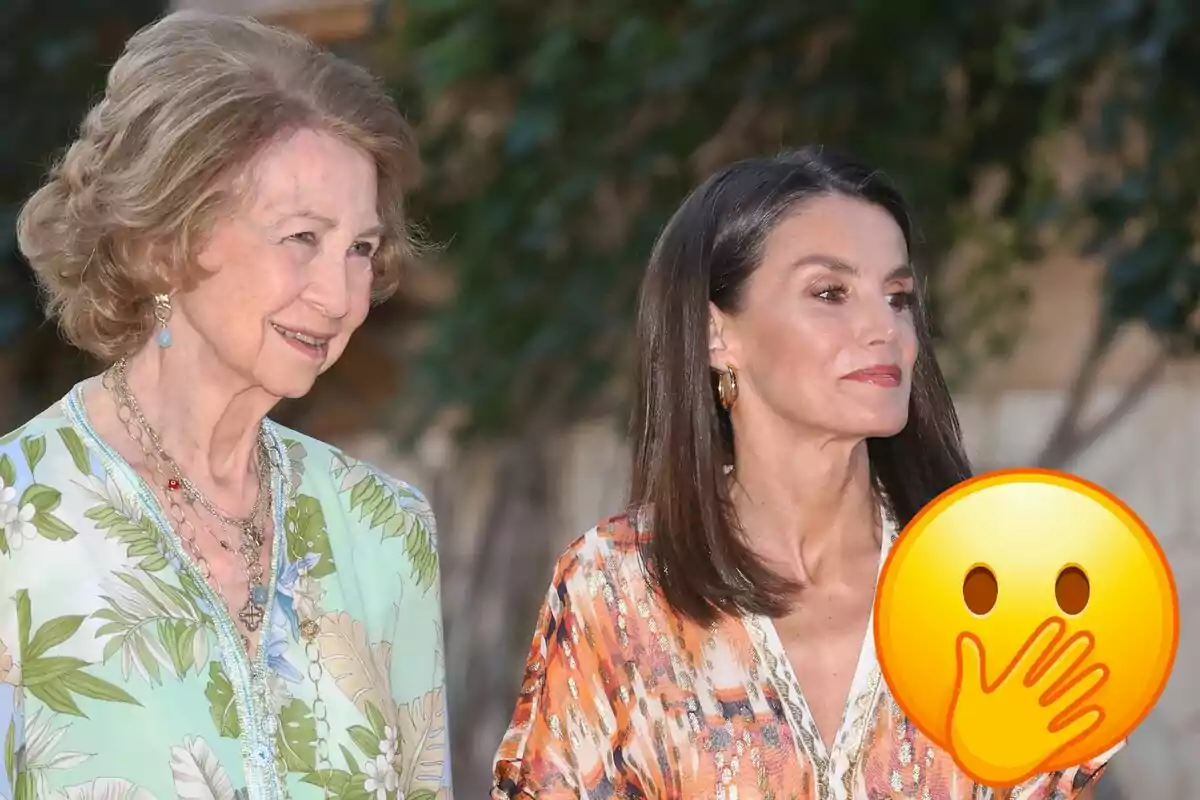 Reina Sofía y Letizia sonríen mientras están de pie al aire libre, Sofía lleva un vestido con estampado floral y Letizia un vestido con estampado colorido, con un emoji de cara sorprendida cubriendo parte de la imagen.