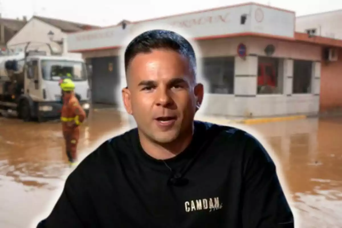 Ángel Gaitán con camiseta negra está en primer plano mientras al fondo se observa una escena de inundación con un camión y un trabajador con uniforme de emergencia.