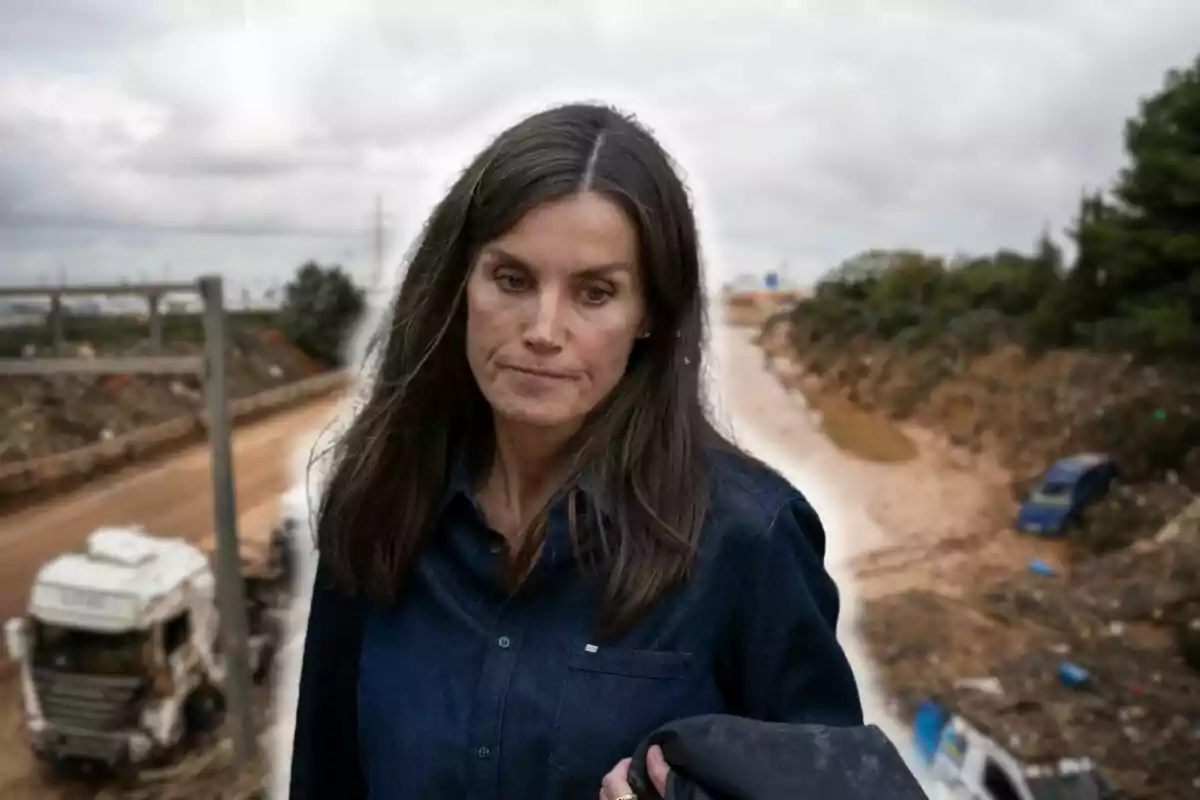 Letizia con expresión seria camina por un área afectada por desastres naturales con vehículos y escombros en el fondo.