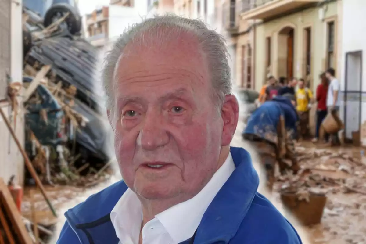 Juan Carlos I con chaqueta azul aparece en primer plano mientras al fondo se observa una calle con escombros y personas trabajando.