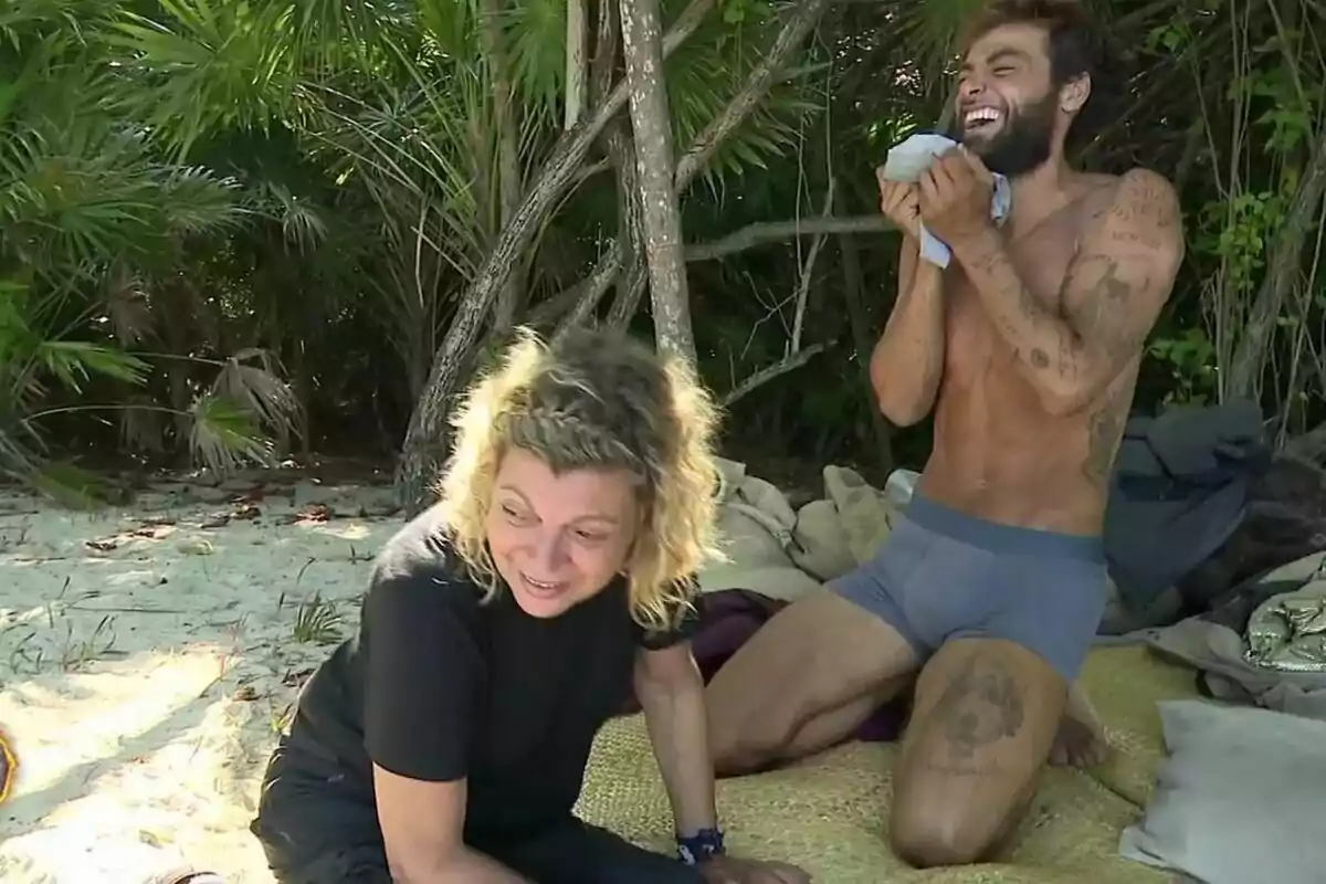 Dos personas en una playa de Supervivientes, Terelu Campos sentada en la arena y Montoya de pie riendo, rodeadas de vegetación.